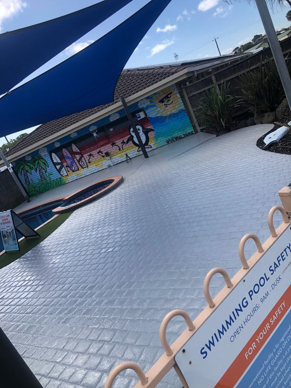 Grey pool surround under blue shade sails, with a swimming pool safety sign